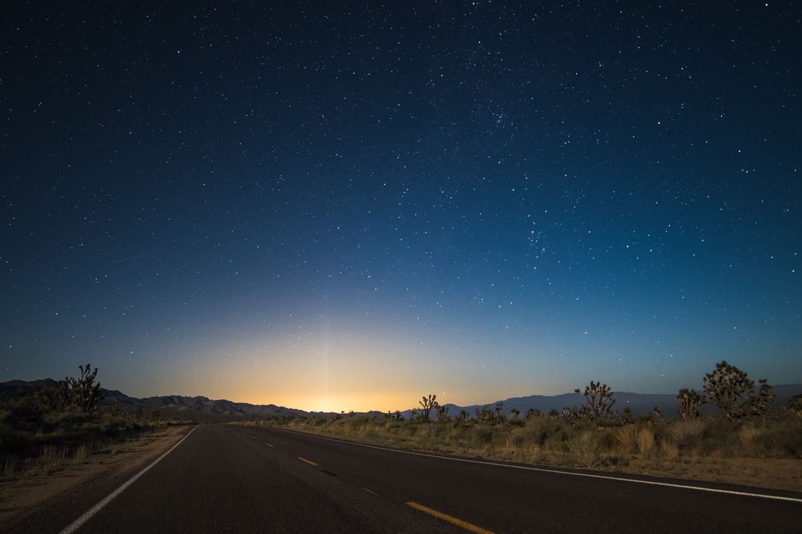 asphalt road in the middle of a desert at night with stars and a bright light emmenating from the horizon